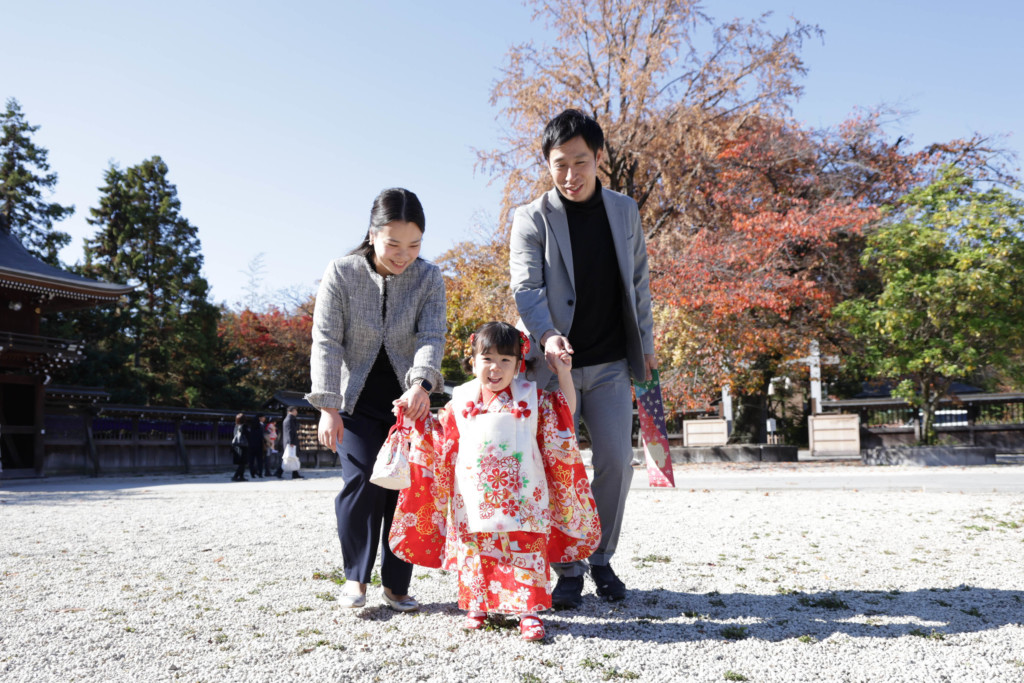 青空と紅葉を背景に家族三人がカメラに向かって歩く七五三写真（立川諏訪神社）