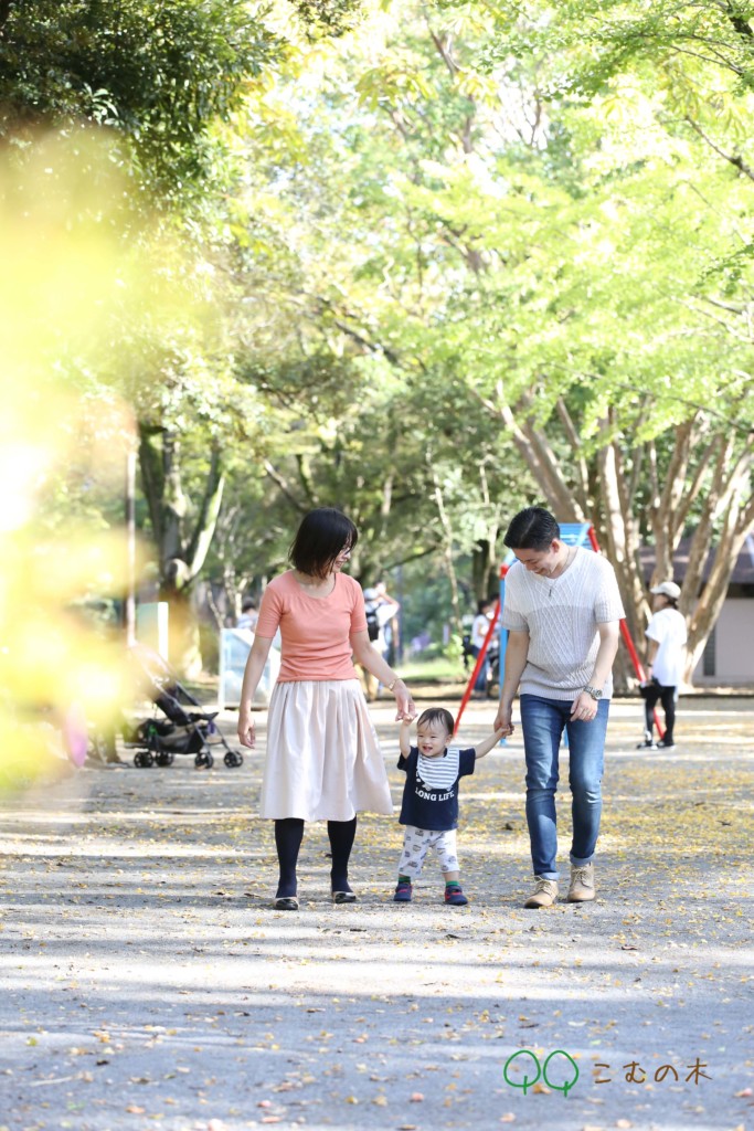 黄色い葉が舞う秋の公園を、歩き始めたばかりの赤ちゃんと両親が手をつないで笑いながら歩く様子。落ち葉の上に柔らかな光が差し込む家族写真。