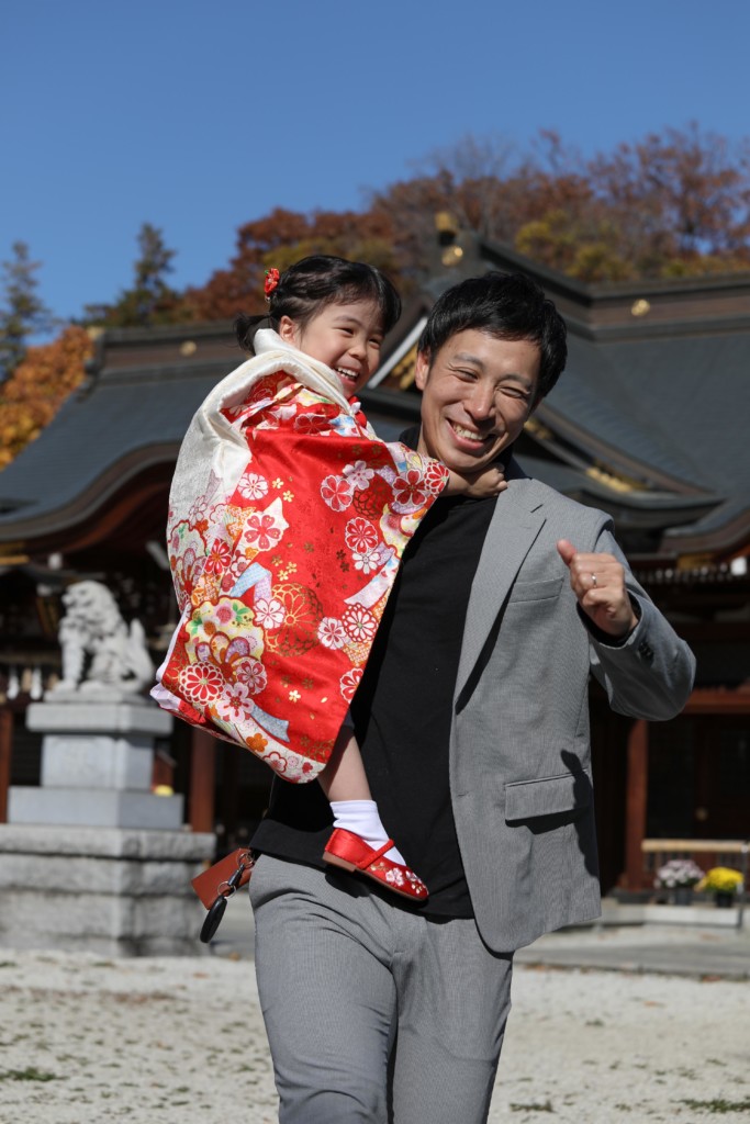 パパに抱き上げられてご機嫌な3歳のお嬢さんの七五三写真（青空と本殿背景）