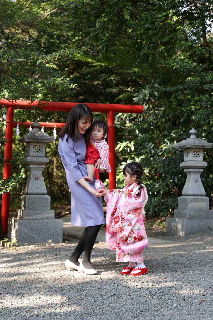 谷保天満宮の赤い鳥居の前で、3歳の女の子がママにどんぐりを渡している七五三の出張撮影。