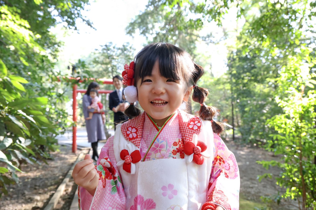 アンパンマンの人形をカメラの上に置き、笑顔を見せてくれた3歳の女の子のアップ。後ろに家族の姿がやわらかく写る。