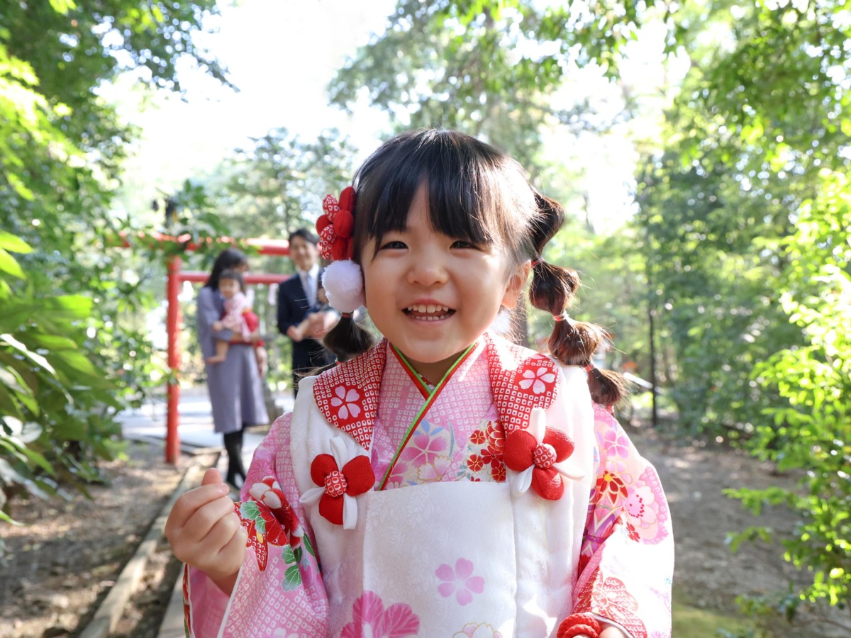 アンパンマンの人形をカメラの上に置き、笑顔を見せてくれた3歳の女の子のアップ。後ろに家族の姿がやわらかく写る。