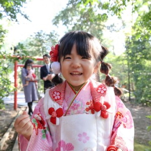 アンパンマンの人形をカメラの上に置き、笑顔を見せてくれた3歳の女の子のアップ。後ろに家族の姿がやわらかく写る。