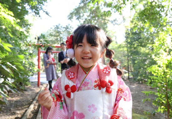 アンパンマンの人形をカメラの上に置き、笑顔を見せてくれた3歳の女の子のアップ。後ろに家族の姿がやわらかく写る。