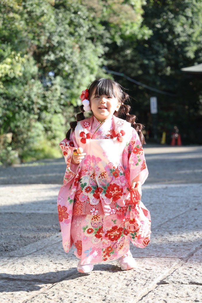 谷保天満宮で七五三の撮影。3歳の女の子が本殿前は少し恥ずかしがり、反対側の緑を背景にしゃがんで撮影した一人写真。